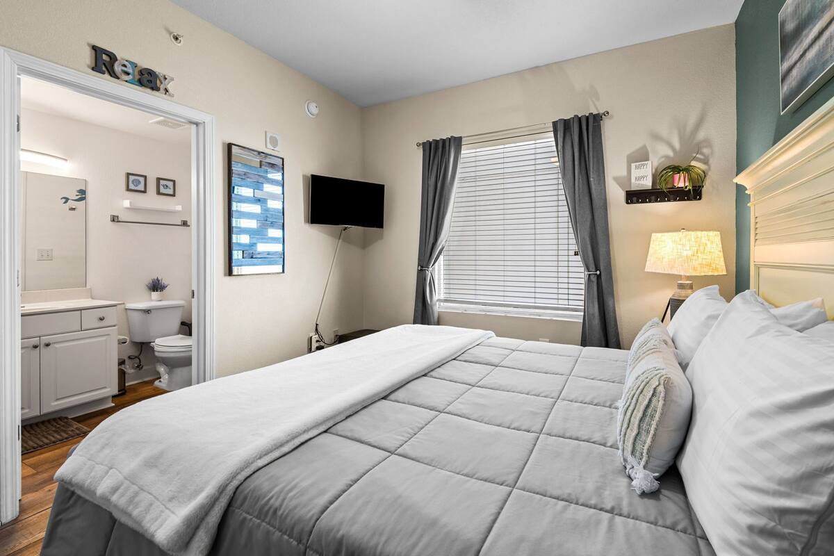 A bedroom with a bed, a television, a window with curtains, a bedside lamp, and a small shelf with a plant.