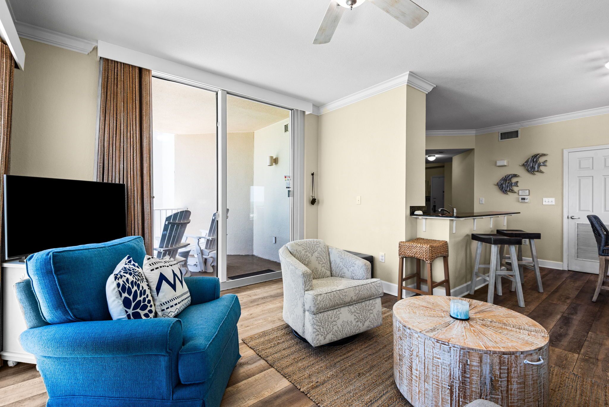 Un salon avec un canapé bleu, un fauteuil à motifs, une table basse ronde en bois, et un coin cuisine à l’arrière-plan.
