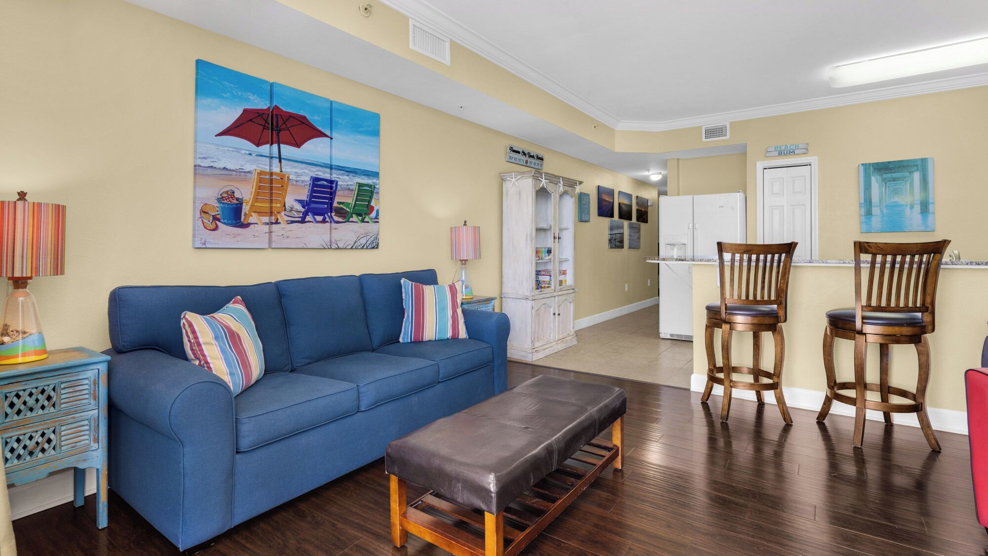 A living room with a blue sofa, a bench, and two wooden chairs. There is a painting of a beach scene on the wall.