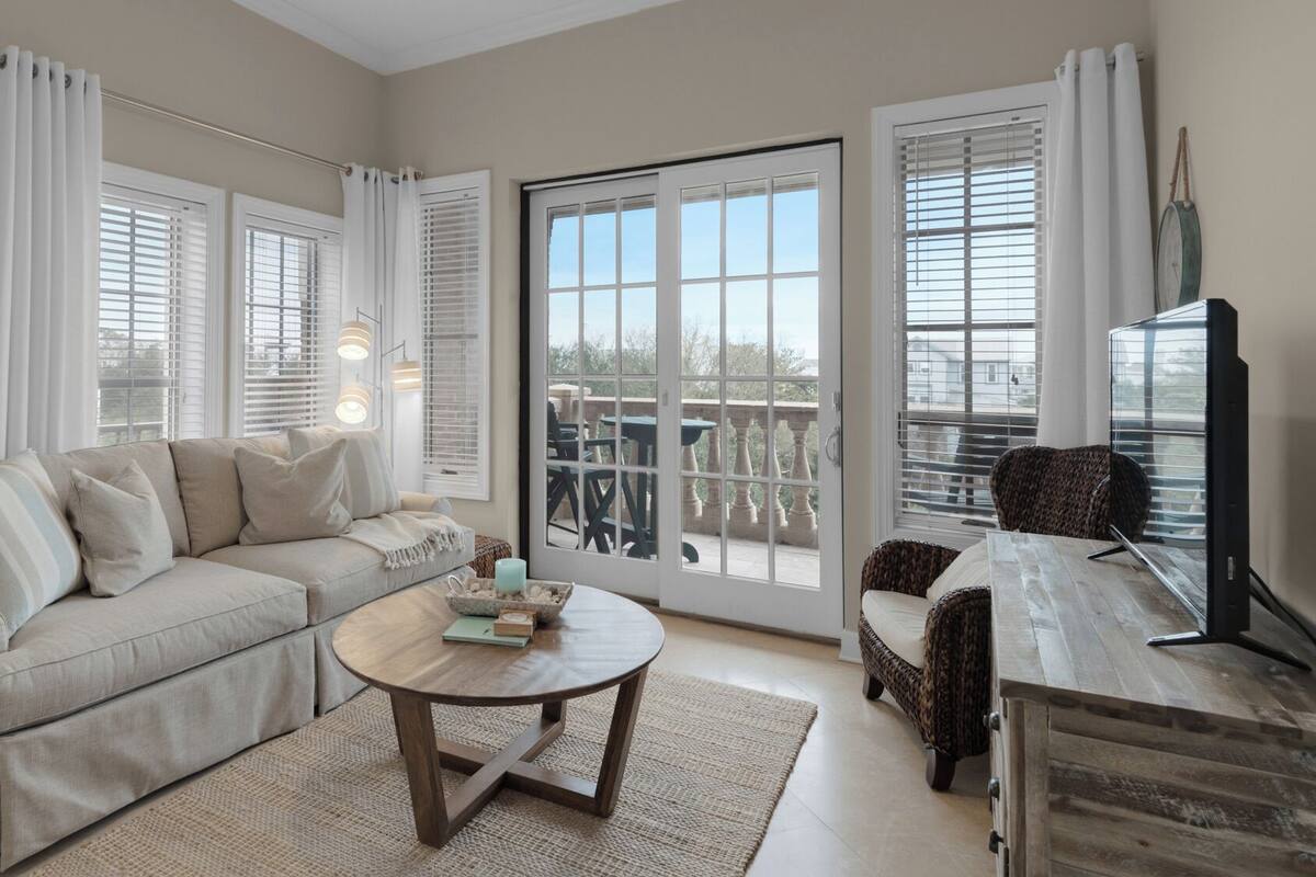 A living room with a beige sofa, a round coffee table, and a wicker chair. There are large windows with white curtains and a view of the outdoors.