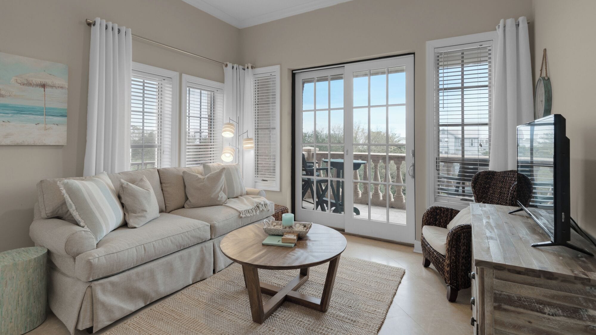 A living room with a beige sofa, a round coffee table, and a wicker chair. There are large windows with white curtains and a view of the outdoors.