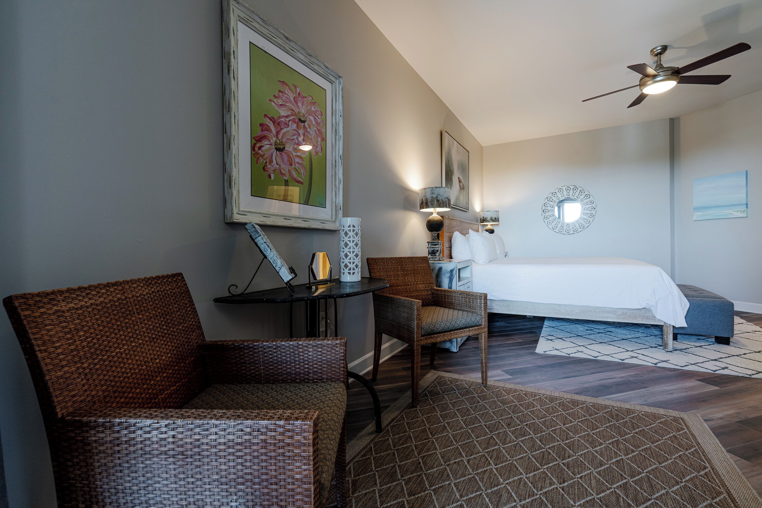 A bedroom with a bed, two wicker chairs, a small table, and a ceiling fan.
