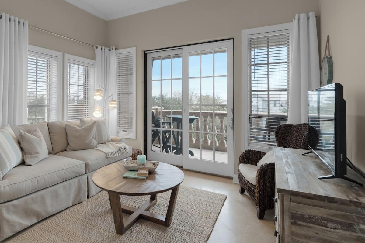 A living room with a beige sofa, a round coffee table, and a wicker chair. There are large windows with white curtains and a view of the outdoors.