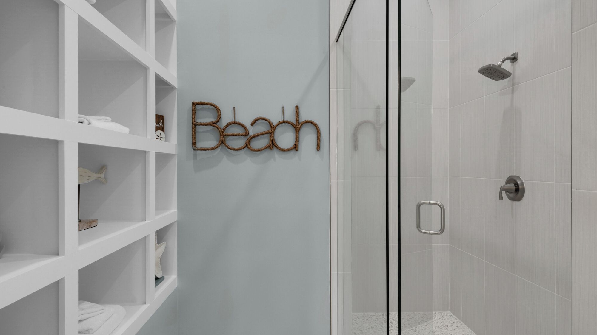 A bathroom with a shower, a white shelf unit, and a wall-mounted sign that reads "Beach".