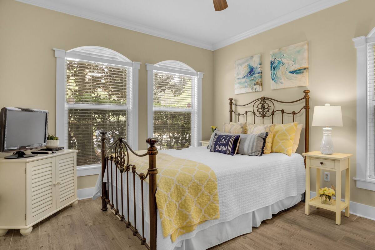A bedroom with a bed, a nightstand, a TV, and large windows with blinds.