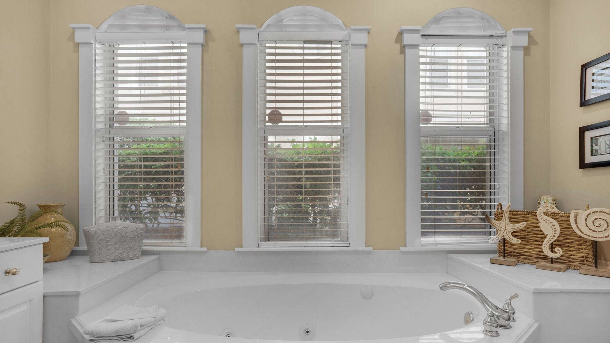 A bathroom with a large bathtub, three windows with white blinds, a sink, and a decorative starfish and seashell arrangement.