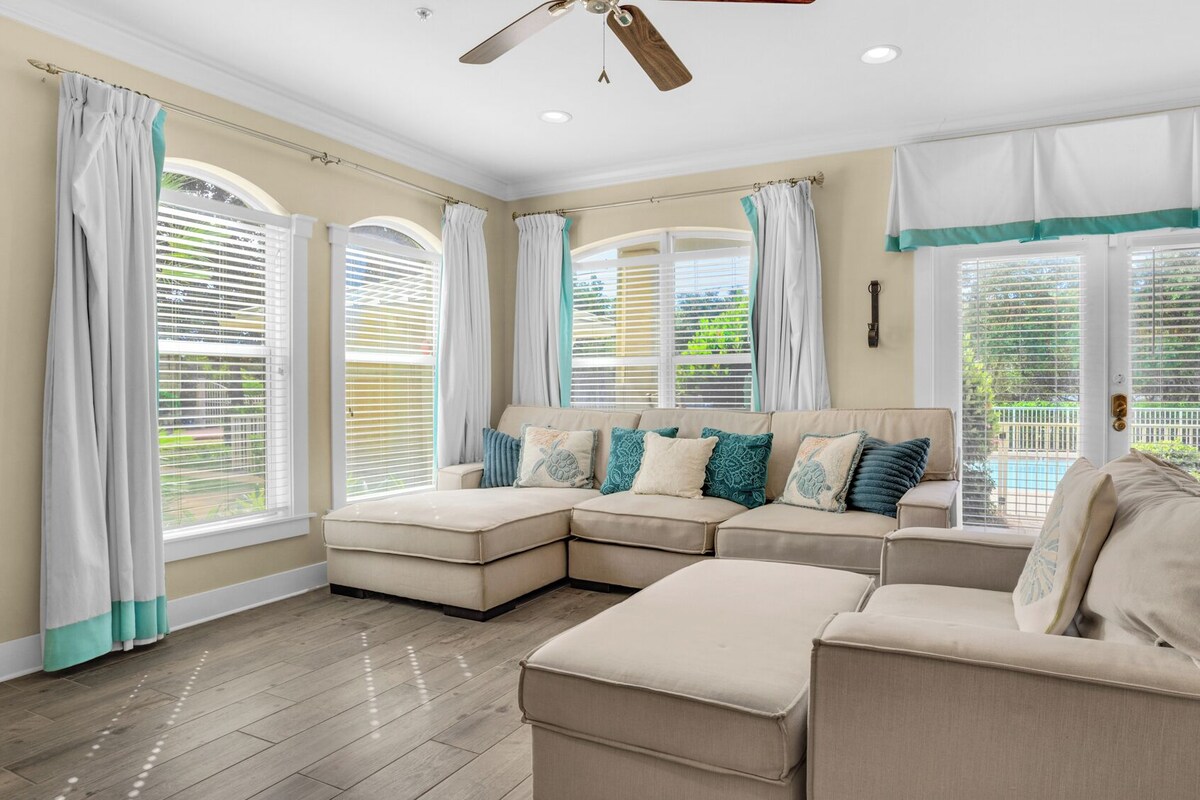 A spacious living room with a sectional sofa, large windows with blinds, and a ceiling fan.