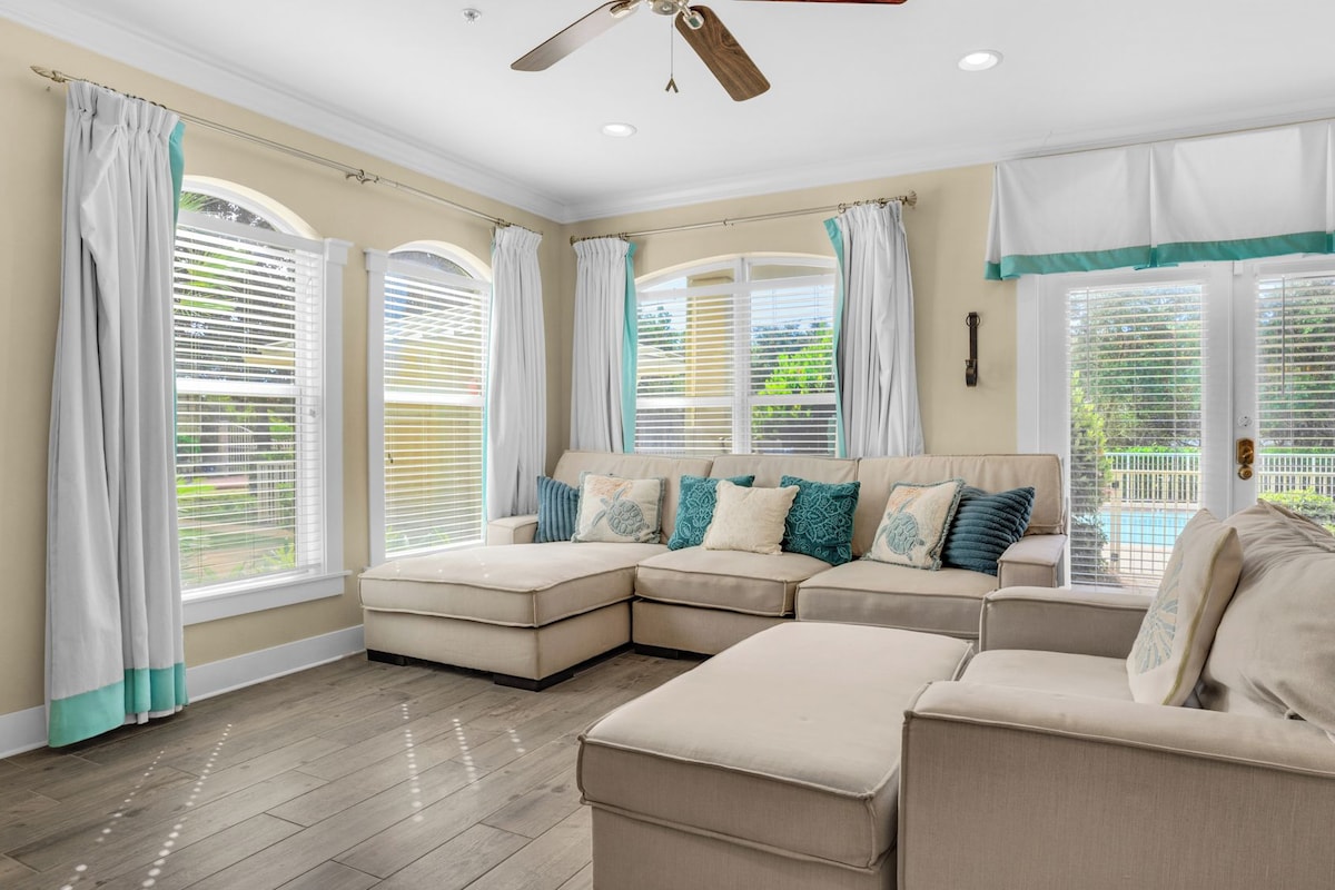 A spacious living room with a sectional sofa, large windows with blinds, and a ceiling fan.