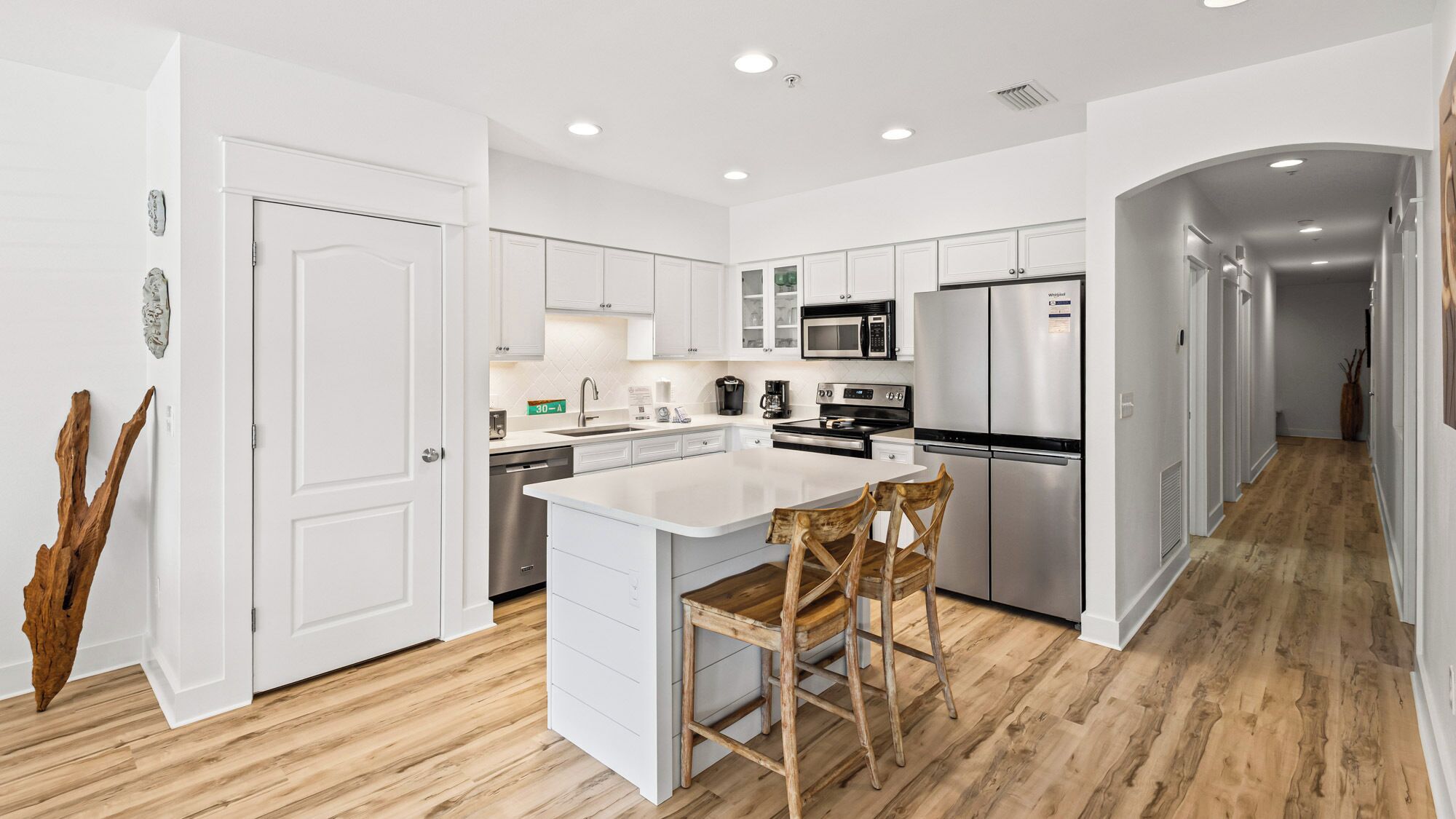 Una cocina moderna con armarios blancos, una isla central, electrodomésticos de acero inoxidable y una silla de madera.