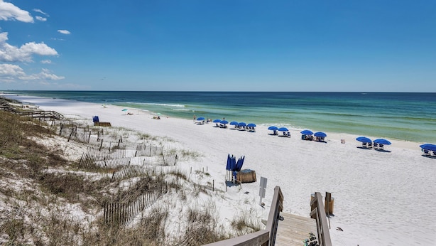 Ein Strand mit weißem Sand, blauen Sonnenschirmen und einer Holzpromenade.