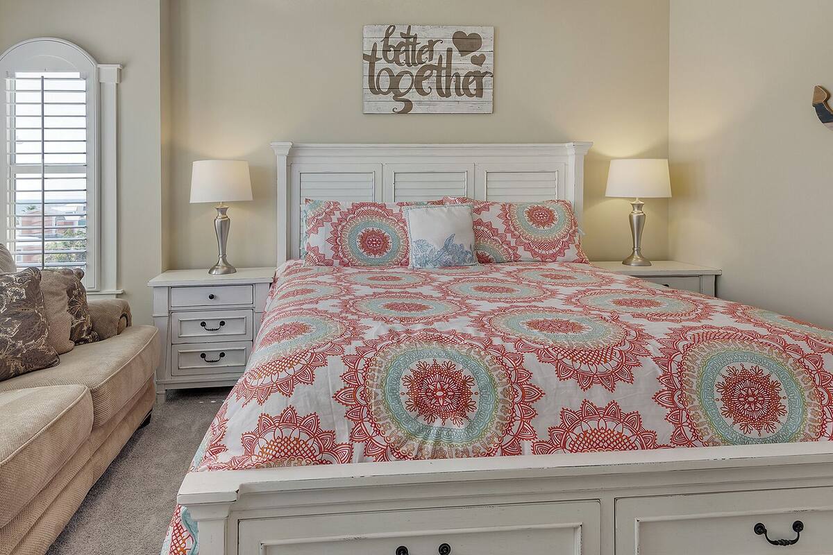 A bedroom with a bed, two bedside tables, a nightstand, a couch, and a window with blinds.