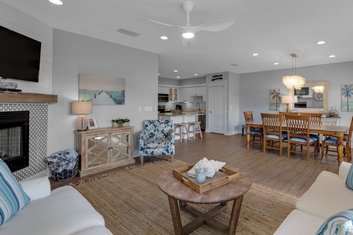 A spacious living room with a fireplace, a dining area with wooden chairs, and a kitchen in the background.