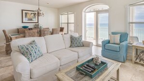 A spacious living room with a white sofa, blue armchair, and a coffee table. There is a dining area with wicker furniture and a chandelier. The room has large windows with a view of the ocean.