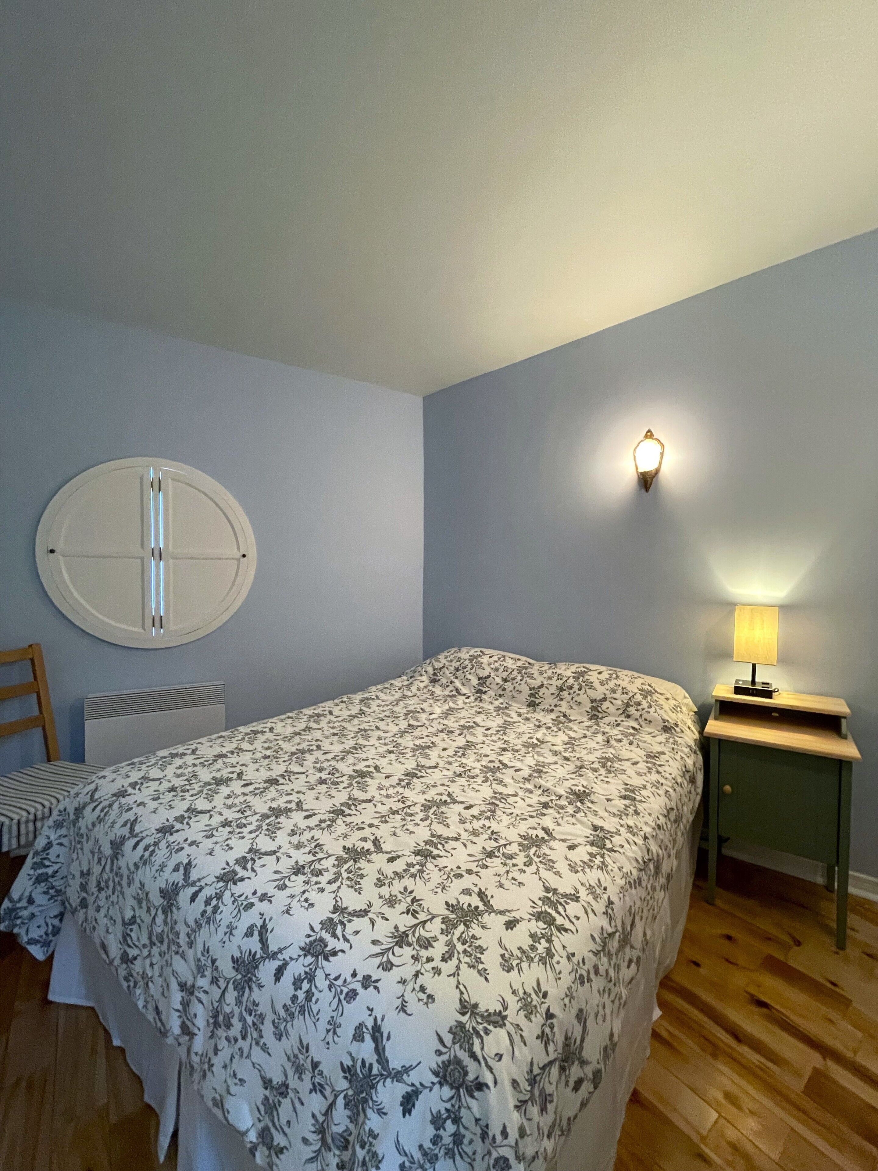 A bedroom with a bed, a nightstand with a lamp, a wall-mounted light, a radiator, and a round mirror.
