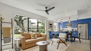 Ein modernes Wohnzimmer mit einem beigen Sofa, einem runden Couchtisch und einem Essbereich mit Stühlen. Im Hintergrund ist eine Küche mit blauen Schränken und Edelstahlgeräten zu sehen.