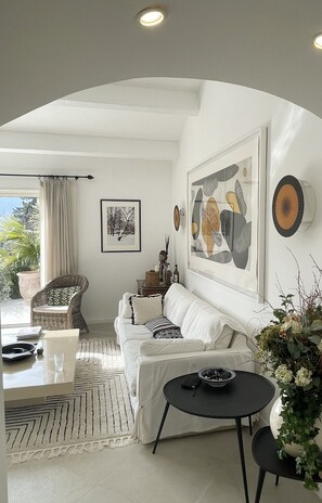 Ein modernes Wohnzimmer mit einem weißen Sofa, einem schwarzen Couchtisch, einem Korbstuhl und einem Esstisch mit Glasplatte.