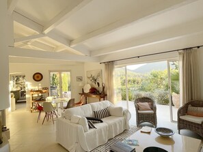 Relax in a spacious living area with a high ceiling and exposed beams, featuring a sofa, wicker chair, and dining area with a balcony offering views of lush greenery and distant mountains.