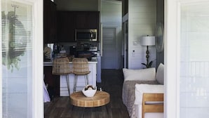 A cozy interior with a wooden floor, a dining area with wicker chairs, a kitchen with dark cabinets and a microwave, and a bedroom with a bed and a lamp. - Riverfront Tiny House- Fishing & Kayaking (Strawberry Plains)