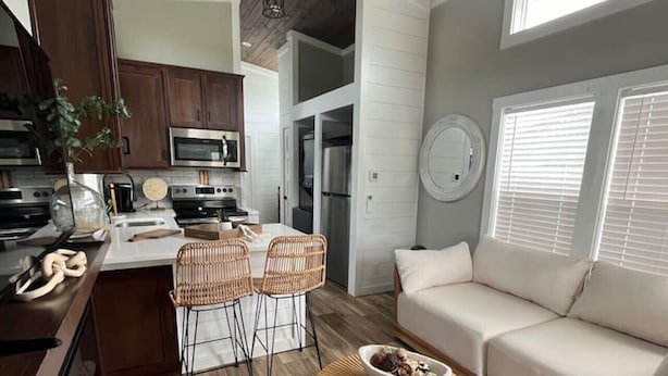 A modern kitchen with wooden cabinets, a dining area with wicker chairs, and a sofa in a living room with large windows. - Riverfront Tiny House- Fishing & Kayaking (Strawberry Plains)