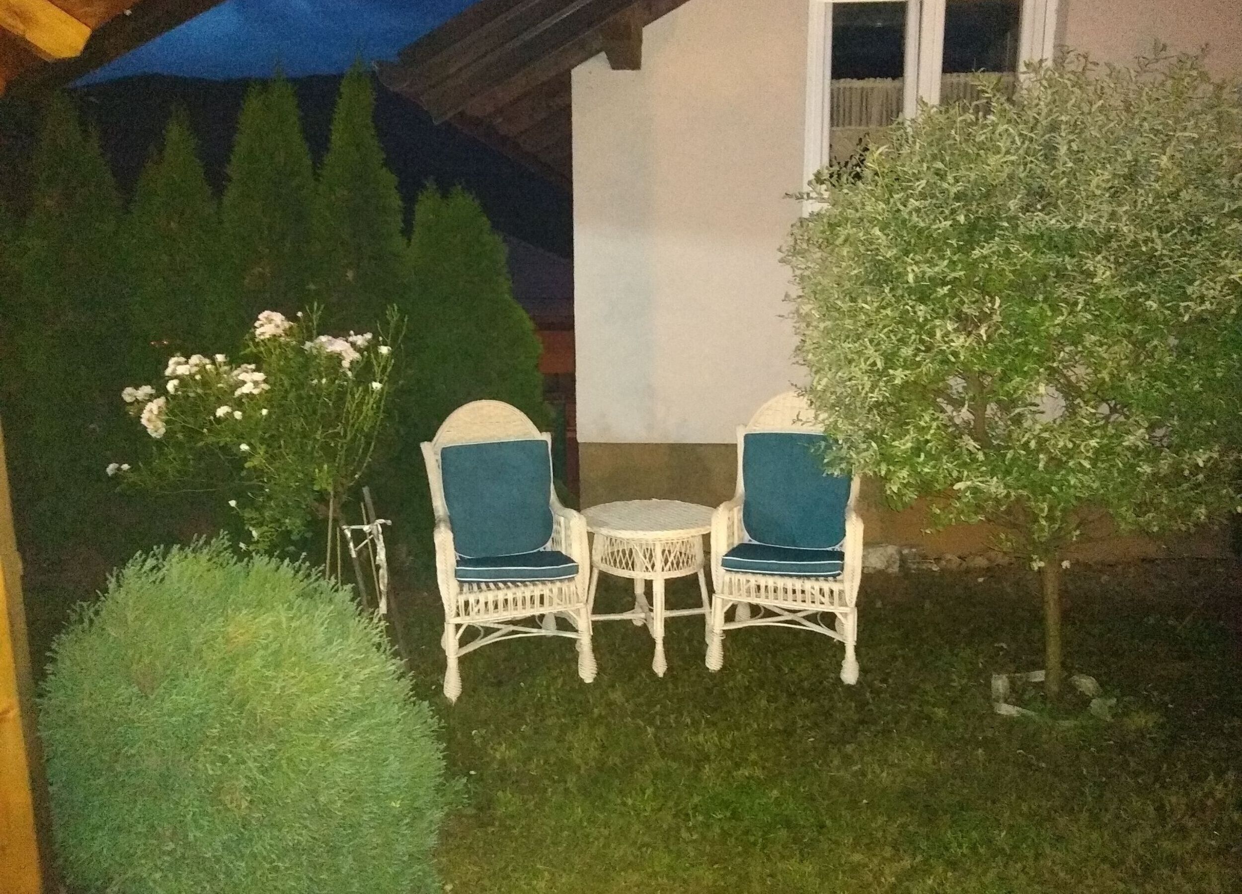 Un área de estar al aire libre con dos sillas de mimbre blanco y una pequeña mesa redonda, rodeada de follaje y un árbol.
