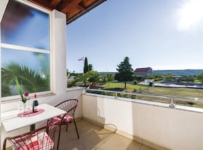 Un balcon avec une table et des chaises, une fenêtre donnant sur un drapeau, et un espace verdoyant avec des arbres et un bâtiment en arrière-plan.
