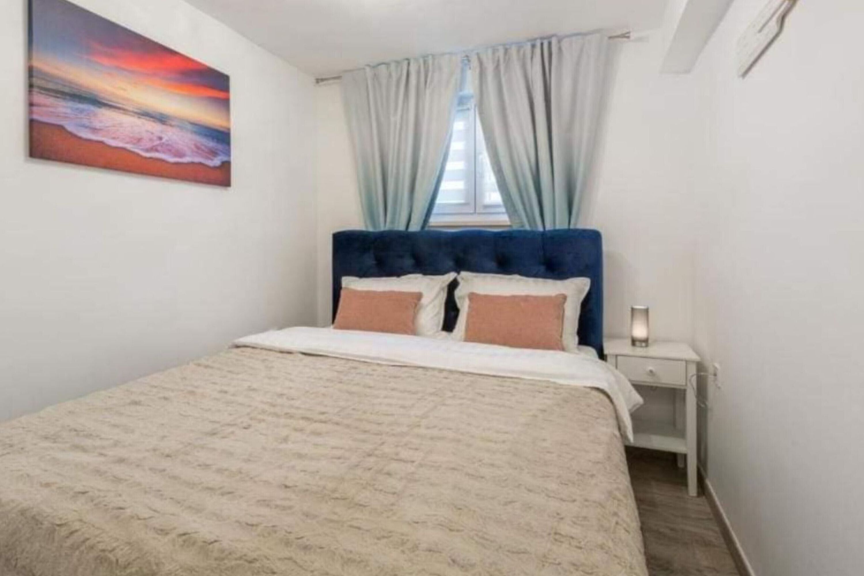 Een slaapkamer met een bed, nachtkastje, schilderij en een raam met gordijnen.