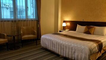 A hotel room with a large bed, a chair, a small table, and a nightstand with a lamp.