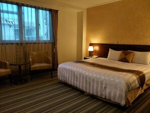 A hotel room with a large bed, a chair, a small table, and a nightstand with a lamp.