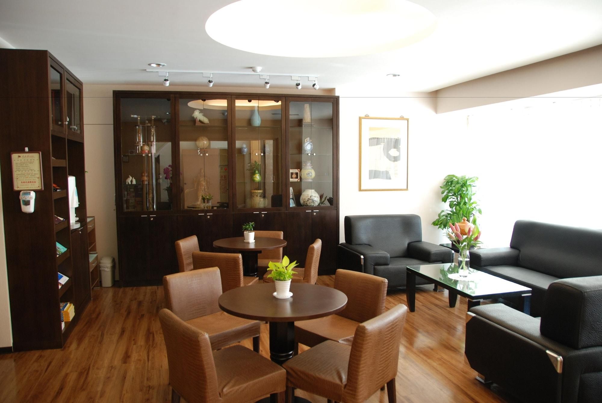 Een hotellobby met een houten vloer, een receptiebalie, een zithoek met stoelen en banken, en een grote kast met glazen deuren.