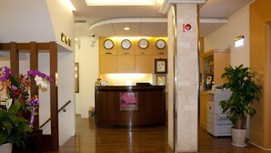 Uma área de recepção de hotel com um balcão de madeira, vários relógios na parede e um lance de escadas com corrimão que leva a outro andar.