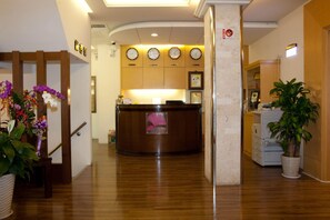 A hotel reception area with a wooden reception desk, multiple clocks on the wall, and a staircase with a handrail leading to another floor. - Hong Cheng Sing Business Hotel (Tainan)