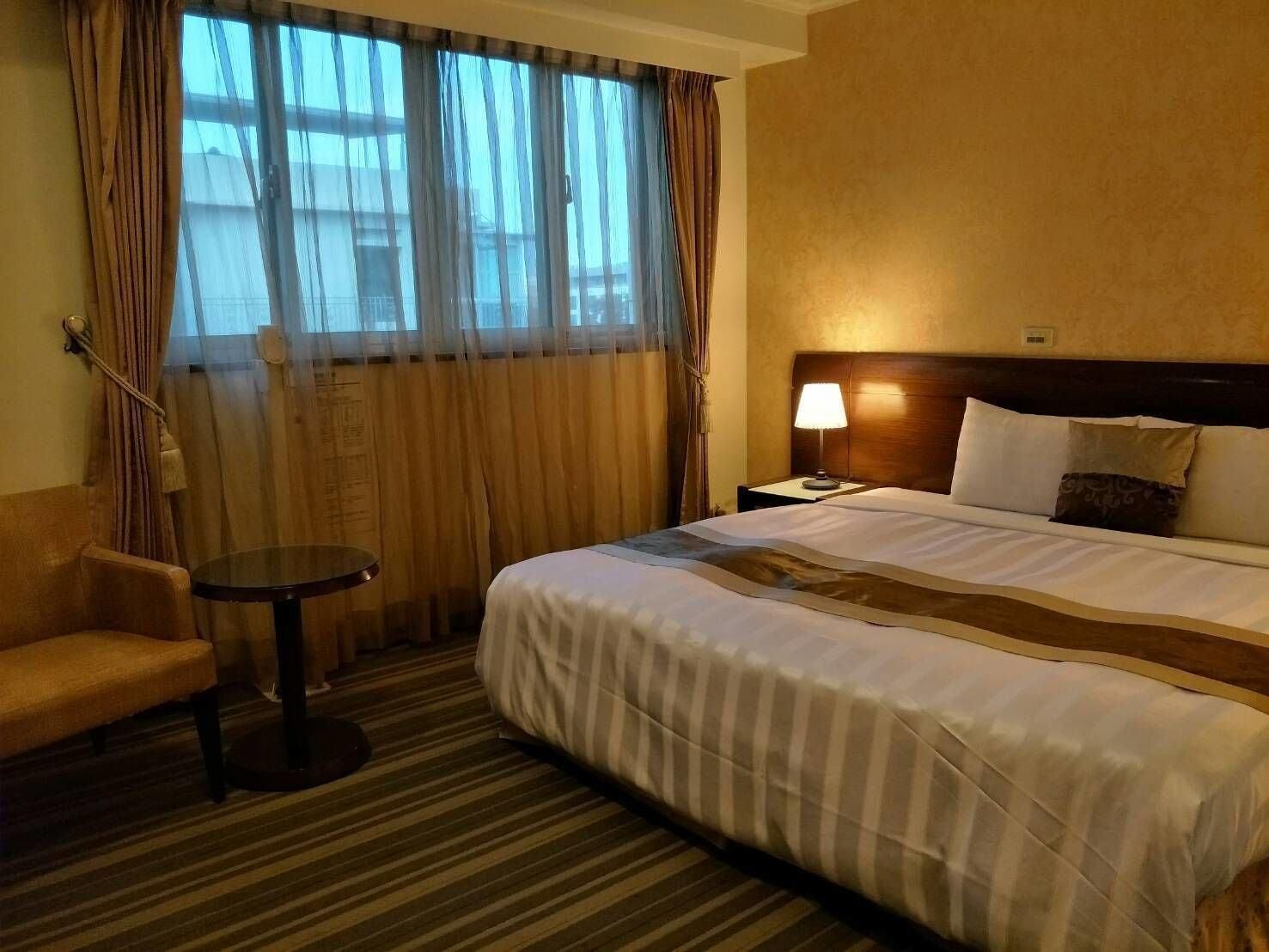 A hotel room with a large bed, a small round table, a chair, and a window with curtains.