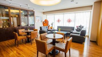 A hotel lobby with wooden flooring, a bar area with glass cabinets, a seating area with sofas and chairs, and large windows with sheer curtains.