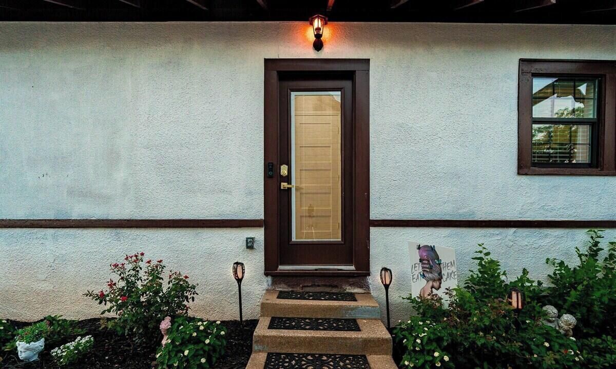 A house with a brown door, a window with blinds, and a small garden with flowers and a stepping stone pathway.