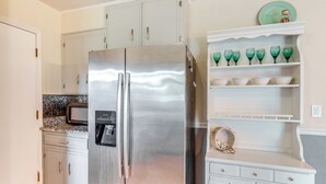 Una cocina con armarios blancos, un refrigerador de acero inoxidable, un microondas y una estantería con vasos y cuencos verdes.