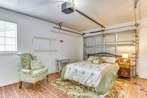 Un dormitorio con cama, una silla, una mesita de noche, una lámpara y un proyector en el techo.