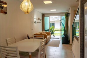 A dining area with a table and chairs, a sofa, a hanging light fixture, and a view of the outside. - Cond. Azul Mar Porto de Galinhas (Ipojuca)