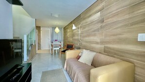 A modern living room with a sofa, a TV, a dining area with a table and chairs, and a wooden accent wall. - Cond. Azul Mar Porto de Galinhas (Ipojuca)