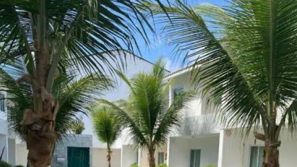 A swimming pool with a wooden deck, surrounded by palm trees and a white building with a green door. - Cond. Azul Mar Porto de Galinhas (Ipojuca)