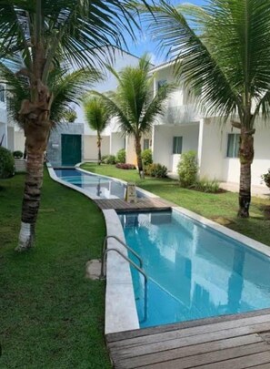 A swimming pool with a wooden deck, surrounded by palm trees and a white building with a green door. - Cond. Azul Mar Porto de Galinhas (Ipojuca)