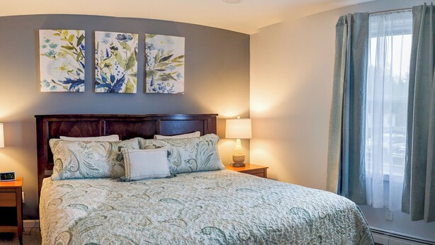 A hotel room with a large bed, two wall-mounted paintings, a nightstand with a lamp, and a window with curtains. - Peaceful Woodland Apt C_Acadia National Park (Seal Cove)