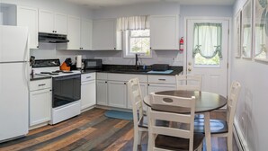 A compact kitchen with white cabinets, a stove, microwave, and a dining area with a table and chairs. - Peaceful Woodland Apt C_Acadia National Park (Seal Cove)