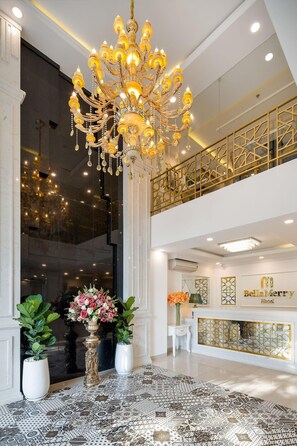 Een statig kroonluchter in een hotel lobby met een hoge plafond, een balkon in dubbele hoogte en een receptiebalie met een bordje.