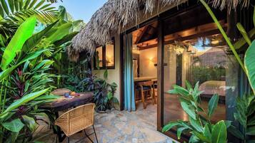 Un área de comedor al aire libre de estilo tropical, con techo de paja, piso de piedra y abundante vegetación.