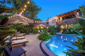 A poolside area with lounge chairs, string lights, and a modern building with a balcony. - Villa Tayrona (Bucerías)