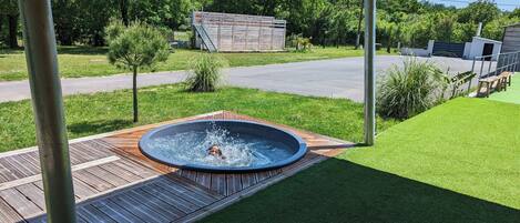 A hot tub in a backyard with a wooden deck, surrounded by grass and trees.
