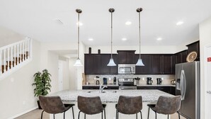 Cocina moderna con armarios oscuros, una isla central con taburetes de bar, electrodomésticos de acero inoxidable e iluminación colgante.