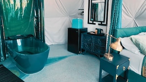 Une chambre moderne avec un lit, une petite table d’appoint, une table de chevet, un meuble noir, un seau vert et une grande fenêtre avec des rideaux.