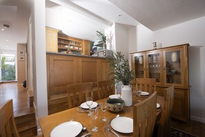 Un coin repas avec une table en bois dressée pour quatre personnes, entourée de chaises en bois. Un meuble avec des portes en verre se trouve à droite, et un vase blanc contenant une plante est posé sur la table.