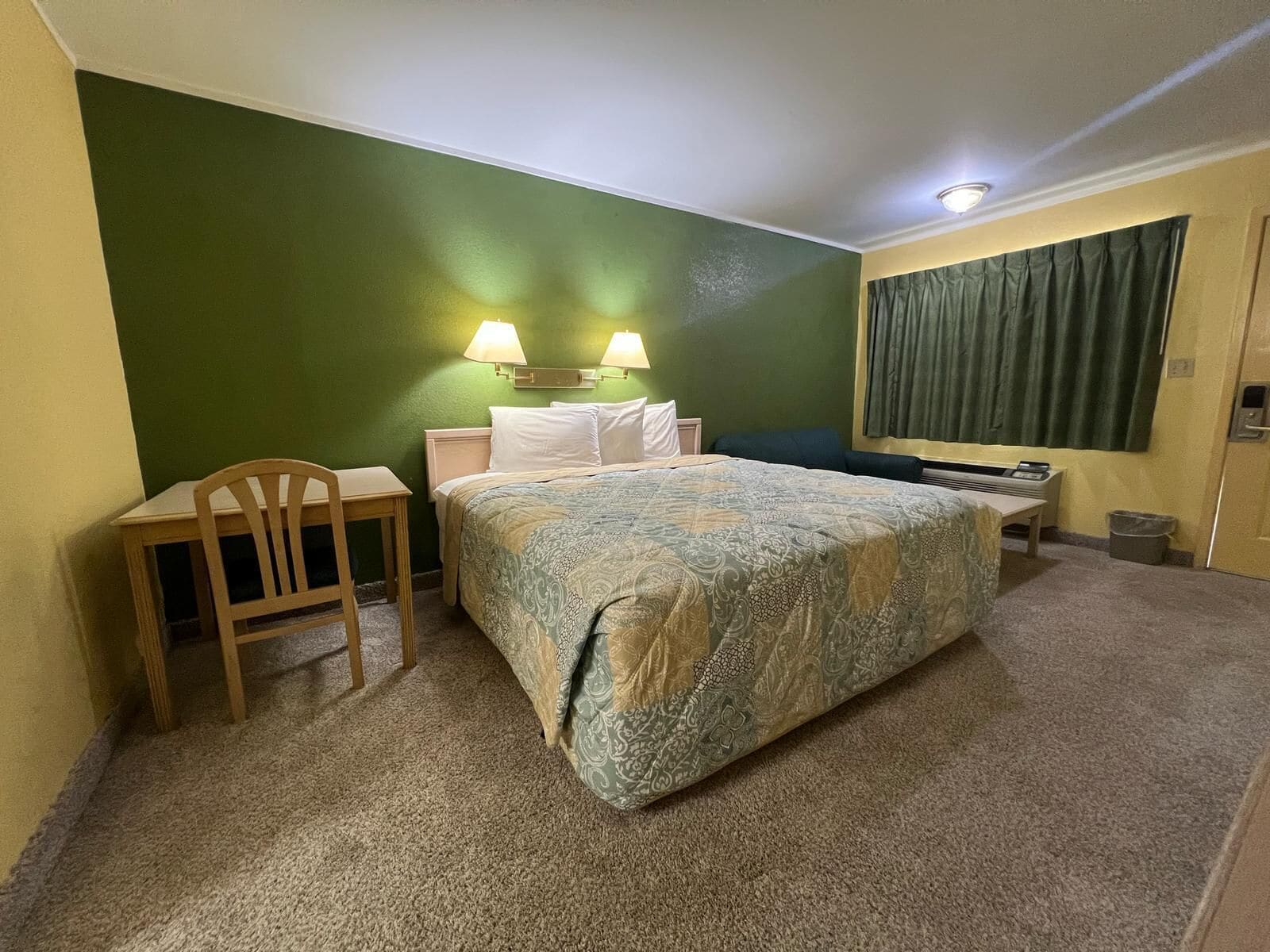 A hotel room with a bed, a desk, a chair, two wall-mounted lamps, and a window with curtains.